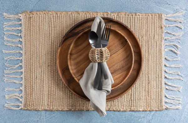 place setting on woven fiber placemat on blue background with two stacked wooden plates and linen napkin with black metal flatware and wicker napkin ring