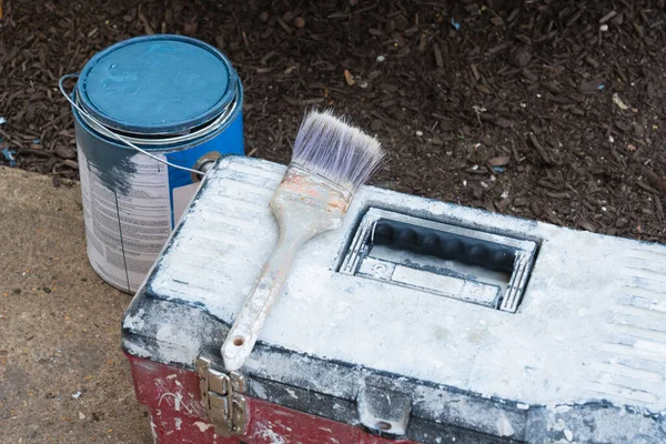 still life of tool kit paintbrush and can of blue paint outside on a sidewalk beside a bed of mulch preparing to paint house trim