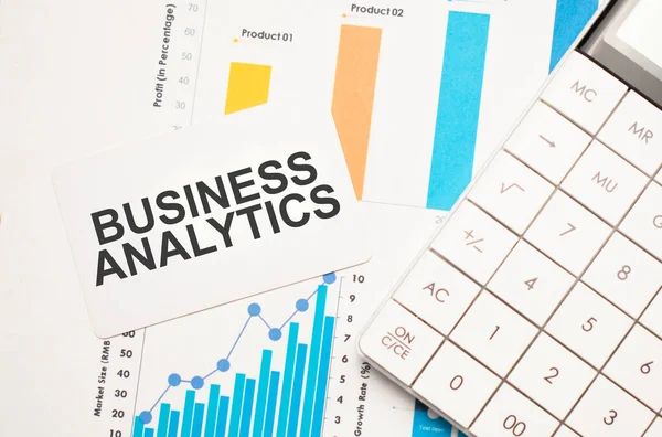 business analytics, text concept. Office workplace table with calculator, graphs, reports and the text business analytics on a small piece of paper on multicolored background.