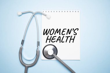 On a light blue table there is a stethoscope, a pen and a sheet of paper with the text WOMEN HEALTH. Medical concept
