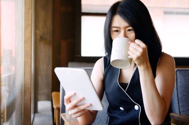 Gülümseyen Asyalı iş kadını bir fincan kahve içiyor ve kafeteryada dinlenmek için sosyal medyada online alışveriş için tablet tutuyor. Tekonoloji konsepti olan iş adamlarının yaşam tarzı.