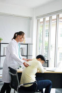 Psikiyatri konusunda uzman olan genç beyaz kadın psikiyatrist, duygusal semptomlar için klinik tedavi gören Asyalı erkek hastanın sırtında stetoskop kullanıyor..