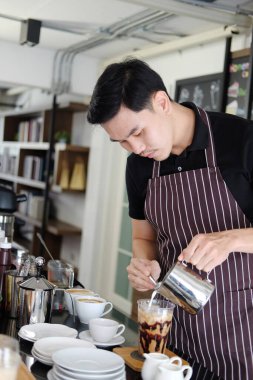 Gülümseyen Asyalı barmen genç adam sabahın köründe modern kafedeki tezgahın siparişine göre plastik bir bardak buzlu kahveye taze süt döküyor..