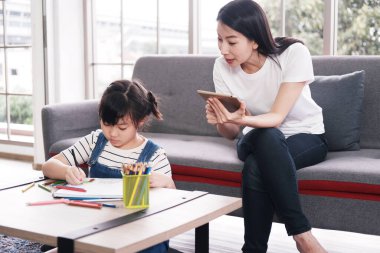 Gülümseyen Asyalı anne ve küçük Asyalı kız çocuk kağıt üzerine ahşap renkli kalemlerle resim yapıyor. Annem oturma odasında tabletle çalışıyor. Ev okulu ve eğitim konsepti