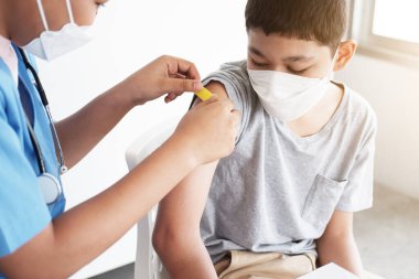 Uzman Afrikalı doktor ya da hemşire kadın, hastanede beyaz bir çocuğa aşı yapıyor ve maske takıyor. COVID-19 'u önlemek için çocuklar için aşılar