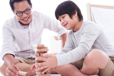 Gülümseyen Asyalı baba ve küçük tatlı çocuk tatilde yatak odasında tahta blok oyunu oynarken eğleniyor ve eğleniyorlar. Aile aktiviteleri ve evde eğitim kavramı için çocuk eğitimi.