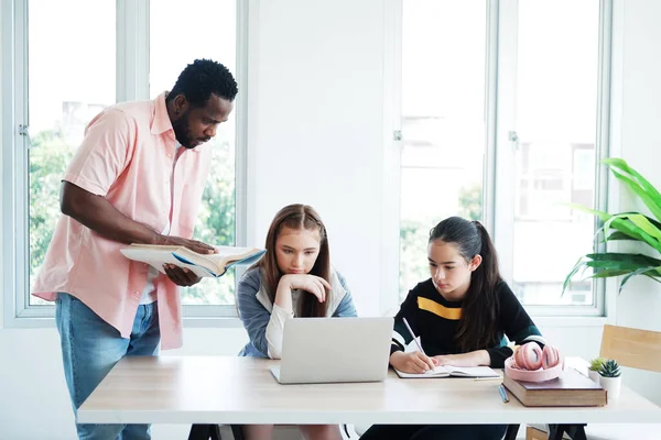 Genç Afrikalı öğretmen ya da öğretmen, sınıftaki beyaz kız öğrencilere internet üzerinden online araştırma hakkında bilgi edinmek için kitap tutuyor ve öğretiyor. Eğitim ve Teknoloji Konsepti.