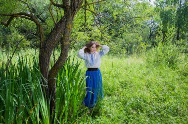 Uzun mavi etekli bir kız ve yeşil çayırlar, bataklık otları ve söğüt ağaçları arasında nakışlı bir gömlek..