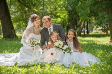 Takım elbiseli damat ve yeşil çimlerin üzerinde bahar bahçesinde uzun beyaz elbiseli gelin ve yaz elbiseli çocuklar. Mutlu bir aile ağaçların arasında güneşli bir günde.