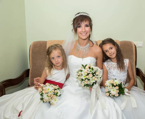Η νύφη και δύο κοριτσάκια με λευκά φορέματα με ένα μπουκέτο λουλούδια κάθονται αγκαλιασμένοι στον καναπέ. — Φωτογραφία Αρχείου