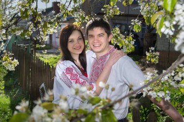 Bahar bahçesinde çiçek açan bir ağacın yanında, beyaz işlemeli gömlekli genç bir çift..