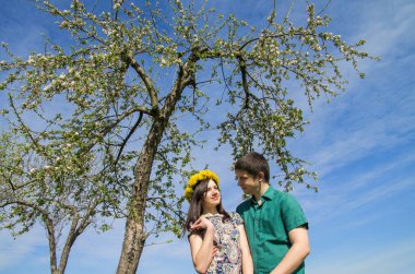 Baharın mavi gökyüzünün arka planında çiçek açan bir ağacın yanında sevgi dolu bir çift. Kafasında karahindiba çelengi olan bir kız..