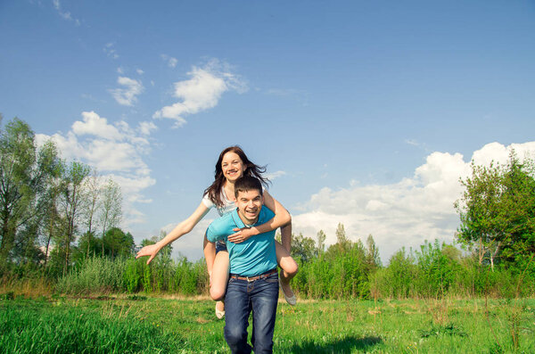 A young man carries his beautiful smiling woman on his back in the park. Couple in love having fun together.