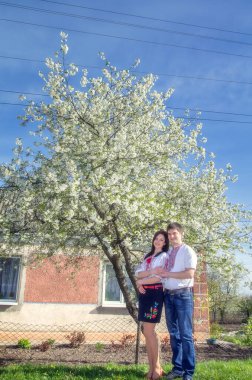 Bahar bahçesinde çiçek açan bir ağacın yanında, beyaz işlemeli gömlekli genç bir çift..