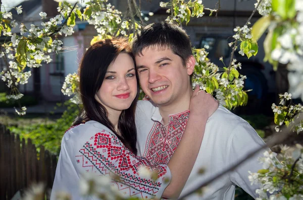在春天的花园里 一对穿着白色绣花衬衫的年轻夫妇站在一棵开花的树上 — 图库照片