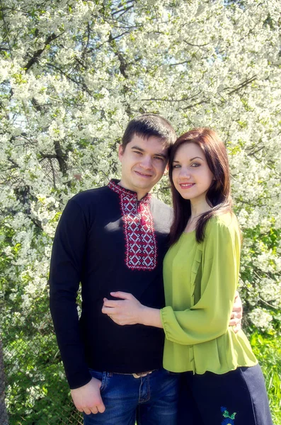 在春天的花园里 一对穿着绣花衬衫的年轻夫妇站在一棵开花的树上 — 图库照片
