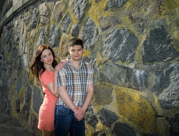 A couple in love near an old stone wall overgrown with moss and lichen. A young girl in a red dress and a young man in jeans.