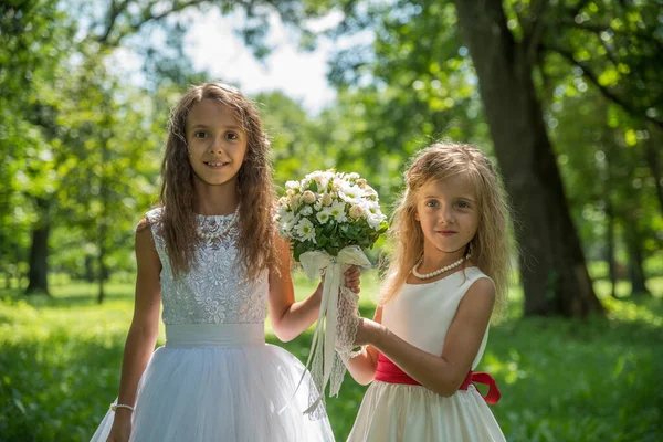 Parkta beyaz elbiseli iki küçük kız kardeş yaz yeşilliklerinin arasında güneşin altında.