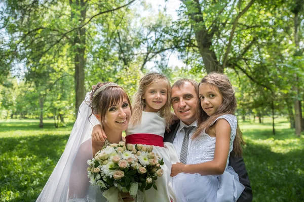 Takım elbiseli damat ve yeşil çimlerin üzerinde bahar bahçesinde uzun beyaz elbiseli gelin ve yaz elbiseli çocuklar. Mutlu bir aile ağaçların arasında güneşli bir günde.