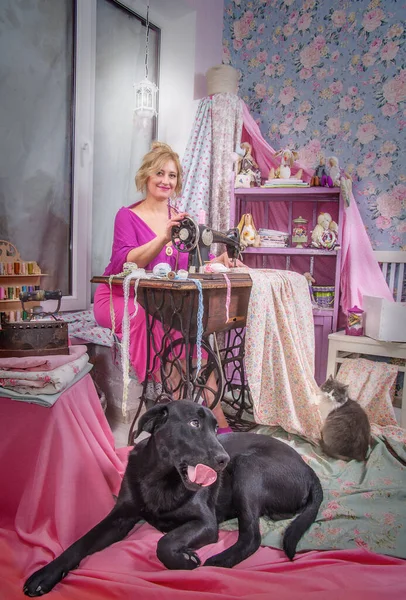 一位女工匠用漂亮的面料在一台老式缝纫机上缝制 附近有一只大黑狗和一只家猫 — 图库照片