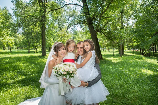 Takım elbiseli damat ve yeşil çimlerin üzerinde bahar bahçesinde uzun beyaz elbiseli gelin ve yaz elbiseli çocuklar. Mutlu bir aile ağaçların arasında güneşli bir günde.
