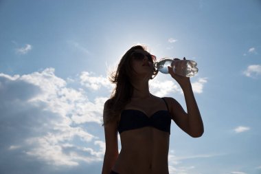 İnce, güzel kız bulutlu bir gökyüzüne karşı arka planda duran bir şişeden su içiyor..