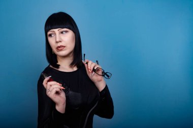 Koyu renk saçlı ve gözlüklü güzel bir kız mavi arka planda elektronik sigara içiyor.
