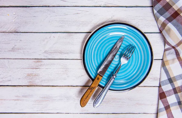 Top view φόντο με άδειο μπλε κεραμικό πιάτο, αντίκες ασημένια μαχαιροπήρουνα σε vintage ξεπερασμένο λευκό ξύλινα διοικητικά συμβούλια. Αντιγραφή χώρου. Καφέ καρό πετσέτα κουζίνας δεξιά γωνία. — Φωτογραφία Αρχείου