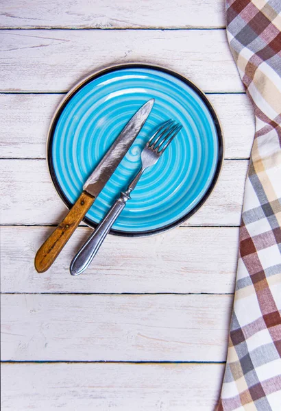 Top view φόντο με άδειο μπλε κεραμικό πιάτο, αντίκες ασημένια μαχαιροπήρουνα σε vintage ξεπερασμένο λευκό ξύλινα διοικητικά συμβούλια. Αντιγραφή χώρου. Καφέ καρό πετσέτα κουζίνας δεξιά γωνία. Κάθετη φωτογραφία. — Φωτογραφία Αρχείου