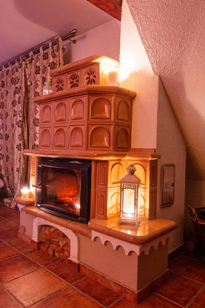 Beiger Kachelofen im alten Stil. Keramische Fliesen in einem Ferienhaus, im Vintage-Stil eingerichtet. Romantisches Nachtbild mit Kamin. Blick aus dem Winkel. Stockbild