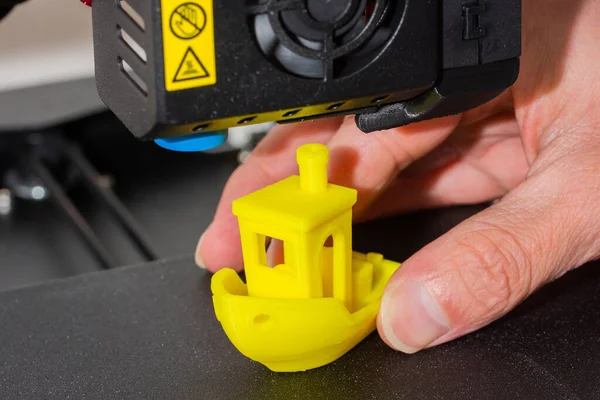 Detailní záběr mužských rukou při dokončeném 3D tištěném modelu žluté PLA lodi. Detail tiskové hlavy s ventilátorem a hotendem. — Stock fotografie