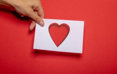 Kadın eli, kırmızı arka planda güçlü bir karton üzerinde aşk sembolü olarak kesilmiş bir kalp şeklindeki kağıt parçasını tutuyor.
