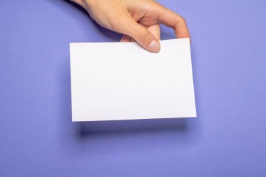 Kadın eli, menekşe renkli arka plan kartonunun önünde fotokopi alanı olan boş bir kağıt parçası tutuyor.