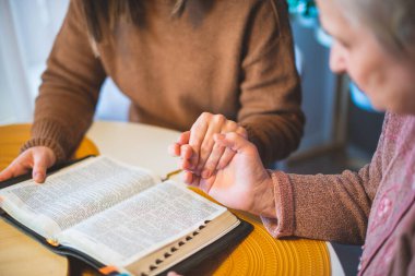 Yaşlı kadın ve kızı el ele tutuşup masada dua ediyorlar.