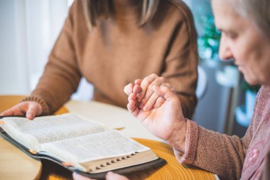 Yaşlı kadın ve kızı el ele tutuşup masada dua ediyorlar.
