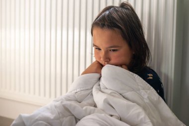 Kendini ısıtmak için radyatörün yanında battaniyeyle oturan genç kız.