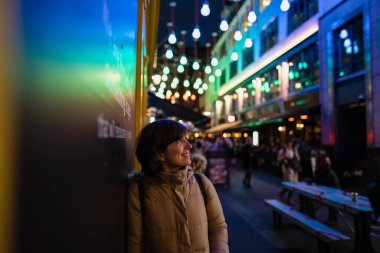 Attractive young female wearing a coat, smiling and looking to one side while being portrayed on the street with Christmas lights in the background.