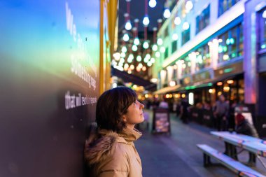 Attractive young female wearing a coat, smiling and looking to one side while being portrayed on the street with Christmas lights in the background.