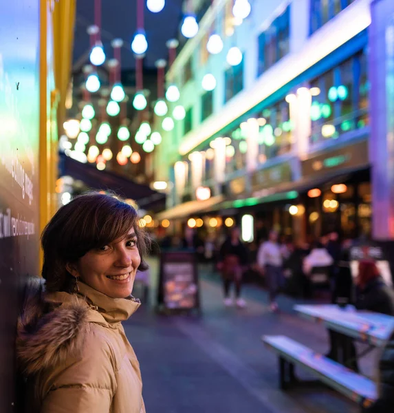 Attractive young female wearing a coat, smiling and posing on the street while being portrayed with Christmas lights in the background.