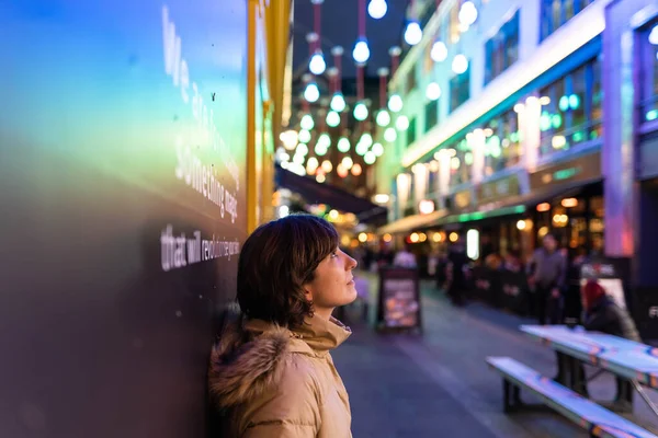Attractive young female wearing a coat, smiling and looking to one side while being portrayed on the street with Christmas lights in the background.