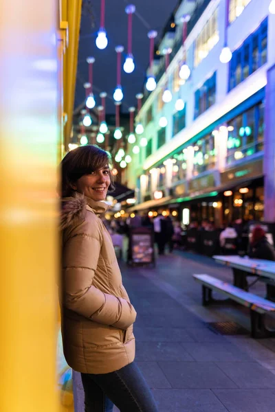 Attractive young female wearing a coat, smiling and posing on the street while being portrayed with Christmas lights in the background.
