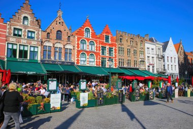 Brugge, Belçika - 9 Ekim. 2021: Yoğun kafeler ve restoranlarla pazar meydanına bakan insanlar, açık mavi sonbahar gökyüzünün dışında oturuyorlar