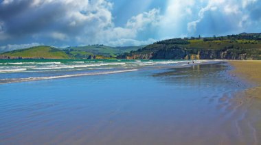 Dalgaların çekildiği kumlu güzel bir akşam manzarası, yeşil tepeler, fırtına bulutları, güneş ışınları - Warrington, Yeni Zelanda