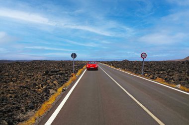 Lanzarote - Timanfaya NP: Uçsuz bucaksız asfalt yolda kara lav kayaları arasında uçsuz bucaksız bir yolculukta çorak engebeli arazide ufukta mavi gökyüzü, beyaz bulut, kırmızı bir Cabriolet araba