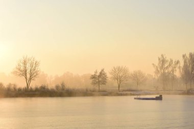 A lake in the winter morning at sunrise, with a small artificial bathing island in the middle, against the fog in the background