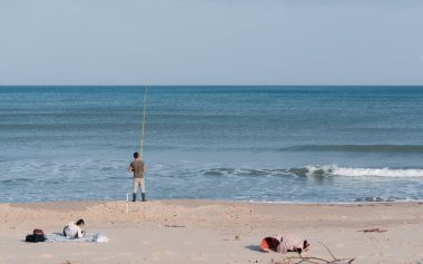 Sahildeki sahne. Bir adam kıyıda duruyor ve denizde balık tutuyor. Sahilde bir kadın yatıyor..