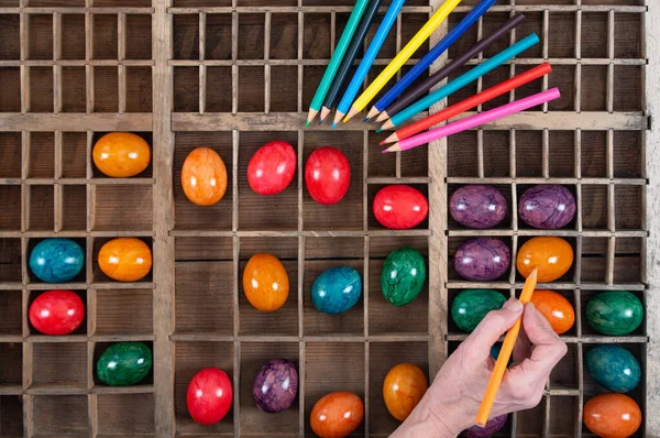 カラフルなイースターエッグを含む多くのコンパートメントを持つ大きな木製の箱の背景と詳細 着色用の色鉛筆が箱の上にあります 手はペンを持ち 卵を描く 卵は光沢がある — ストック写真