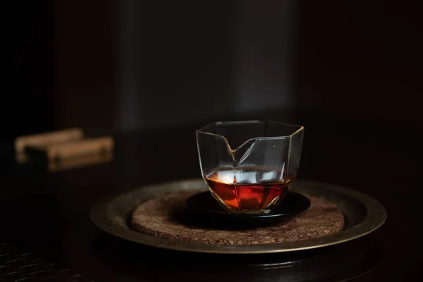 暗い背景を背景に暗いテーブルの上に小さな中国のガラス瓶が立っています 鍋にはキャラメル色のお茶が入っています 鍋は盆に乗っている — ストック写真