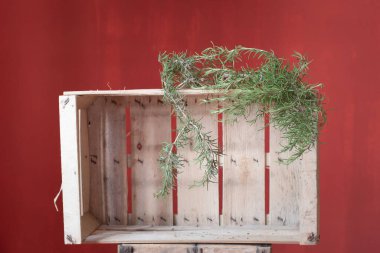An empty rustic wooden box stands against a red background. On the box is a sprig of rosemary to dry