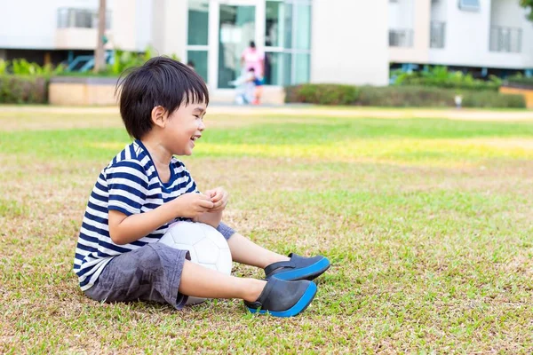 亚洲小男孩一个人坐在公园的草坪上 手里拿着足球 — 图库照片